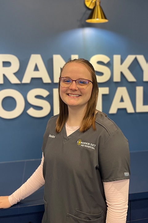 A woman with brown hair and glasses, wearing grey scrubs with a floral logo, smiles warmly in a reception area. The background reveals part of a sign that reads "RAN SKY HOSPITAL" on a blue wall, suggesting she's a dedicated vet ready to assist her furry patients.