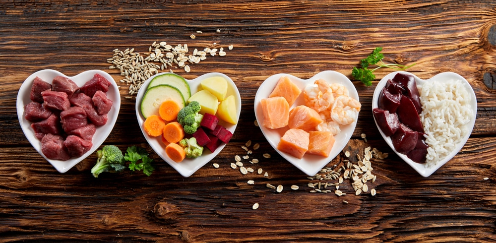 Four heart-shaped bowls on a wooden surface contain enticing treats fit for a pet's delight: raw meat cubes, mixed sliced vegetables, salmon and shrimp pieces, and rice with red meat chunks. Separated by scattered grains and herbs, this setup could impress even the pickiest veterinarian.
